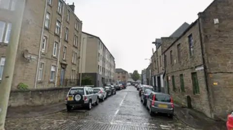 A cobbled street with tenements and cars on either side