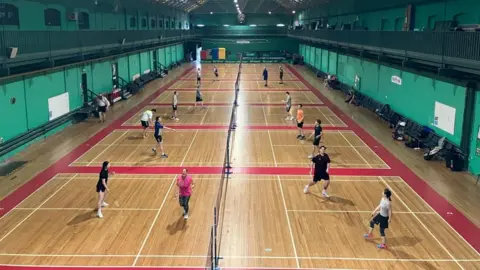BBC A plan of a long gym installed for badminton, with land marked on wooden floors. The mixed genre players are held on each side of the nets, playing two games.