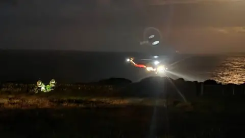 HM Coastguard A dark night with the lights of a helicopter showing over the sea and a few rescuers in yellow jackets standing on shore