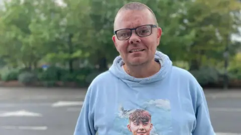 Martin Cosser - a man with short hair and glasses, smiling looking directly in the camera, standing in front of some trees. He is wearing a light blue jumper on with a picture of his son, Charlie Cosser