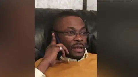 Thames Valley Police Calvin Olawale Babatunde wearing an orange jumper with a white collared shirt and dark-rimmed glasses. He has a goatee and is holding a phone to his ear.