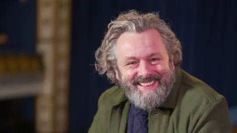 Michael Sheen, who has long grey hair and a beard, smiles to camera while sitting in a theatre 