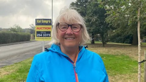 Linda Debres stands in front of a for sale sign