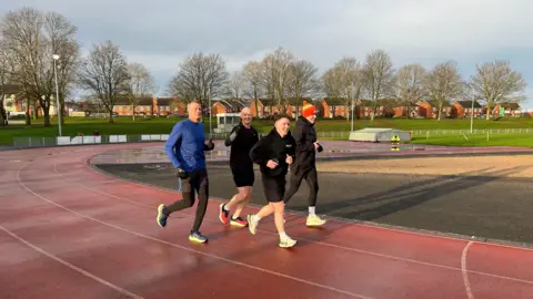 Four people run around the bend of an athletics track, bathed in sunshine. They are mainly wearing black gear, while one has a blue top on and another is wearing a bobble hat.