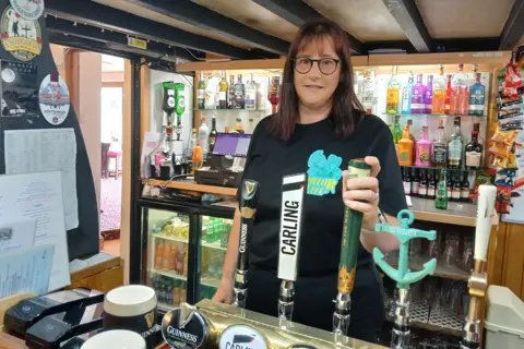 BBC/ALAN WEBBER Sue Maltby stands behind the bar of the Blue Bell Inn. She has shoulder-length, dark brown hair and wears a black T-shirt. She rests a hand on one of five beer pumps. In the background are drinks fridges and rows of spirit bottles.