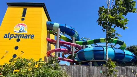 A yellow building with a logo reading Alpamare on the side. Several brightly coloured tube waterslides are exiting the side of the building and weaving around each other.