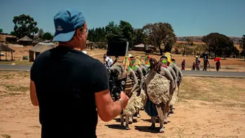 Adam Docker Docker with his back to the camera holding a camera filming people dressed in traditional clothing, they appear to be marching in formation. The filming is taking place in Lesotho, Africa.