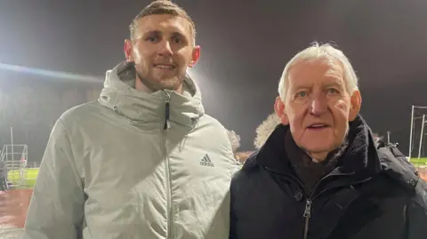 Simon Thake Two men in thick winter jackets stand under the lights at an athletics track. The younger man on the left is taller with fair hair and a beard. The older man on the right has white hair.