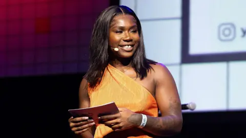 Jade Barnett in an orange dress standing on stage. She holds notecards and wears a microphone. She is at the Youth Music Awards 2024.