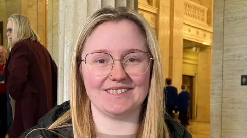 Katie Heagarty-Biggerstaff has long blonde hair, a striped top and black coat.