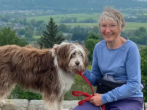 Joan Lyon Joan Lyon, a 75-year-old woman, with grey bobbed hair and glasses, wearing a blue sweatshirt. She is holding a brown and white dog with a red lead.