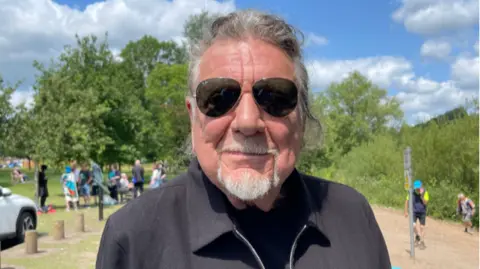 Robert wears a black top and jacket and sunglasses. His hair is tied back.
