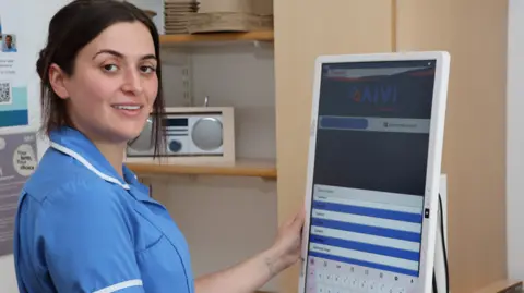 Danielle Jackson is a young woman in her 30s with dark hair tied back wearing a blue nurses uniform. She is holding a large tablet and looking at the camera 