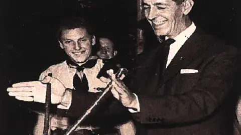 Geoff Pinney Black and white grainy photograph of an older man in a suit and tie, holding two divining sticks in front of him. In he background can be seen a boy wearing a scouts uniform, who is smiling and the face of a second boy behind him