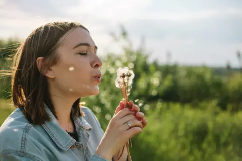 Young woman blowing dandelion seed in meadow outdoors in sunny day. Vibrant summer portrait. - stock photo