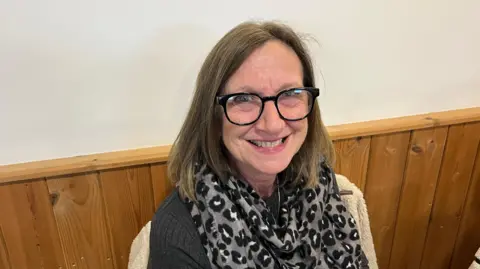 John Devine/BBC Linda Doggett, has brown shoulder length hair parted on the top, she ahs heavy dark framed glasses and a black top with a grey and black chiffon style scarf around her neck, she is sitting in a café with pine wood panelling behind her.