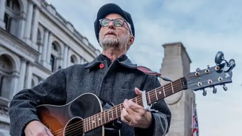 Getty Images Billy Bragg, wearing a black flat cap and glasses, strums an acoustic guitar. He is also wearing a grey woollen coat with a red button. He is looking up, away from the camera, in front of the Cenotaph in Whitehall, London.