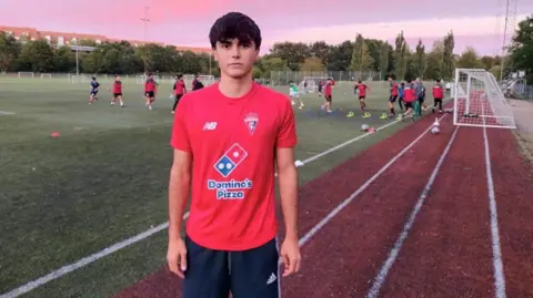 A young man wearing a red T-shirt and tracksuit stands facing the camera with a serious expression on the edge of the football field with a playing team and the sunset sky behind him.