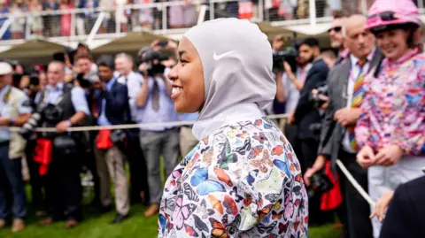 Goodwood Racecourse A woman wearing a hijab and a colourful horse-riding top. There is a crowd in the background. 