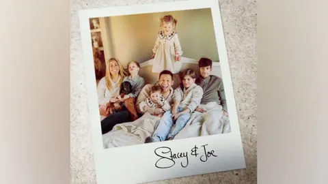 Optomen/Bater & Street Stacey Solomon on the left in her house, sitting with her husband Joe Swash and four of their children who are aged from baby up to 16. They are all wearing neutral colours and smiling at the camera. One girl aged around, four, is standing behind Joe.