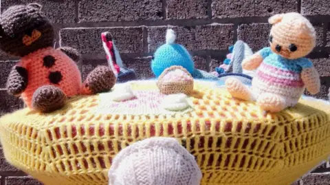 Mail Trail for Hospiscare 2025 A yellow knitted topper with crochet bears having a picnic. There is a seashell on the front. Behind it a brown brick wall.