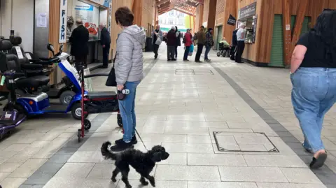Owen Sennitt/BBC A woman with a black dog stands in the middle of Great Yarmouth's market while a woman wearing jeans and a black shirt walks past