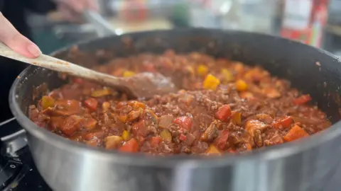 A close up of bolognaise cooking in a large silver pan. The pan is filled with minced beef, red peppers, yellow courgette, onions and a red tomato sauce. There is a wooden spoon balanced on the edge of the pan.