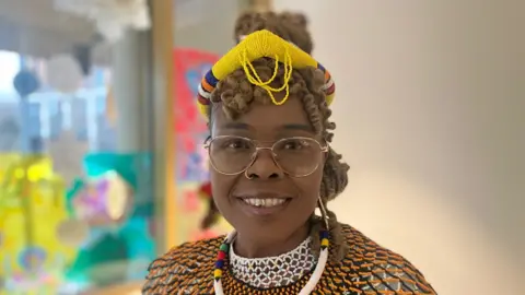 BBC/Elizabeth Baines A woman with gold glasses and traditional South African attire smiles at the camera. Behind her, a blue and yellow window display is blurred.