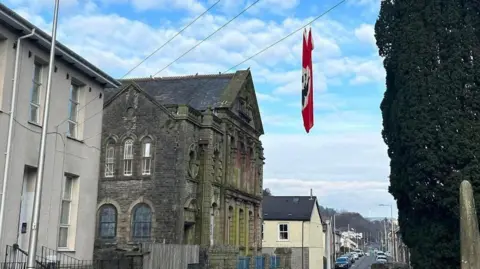 A street in Ton Pentre with wire going across the street. On one of the wires hangs a large red flag, with a white circle and a black swastika,