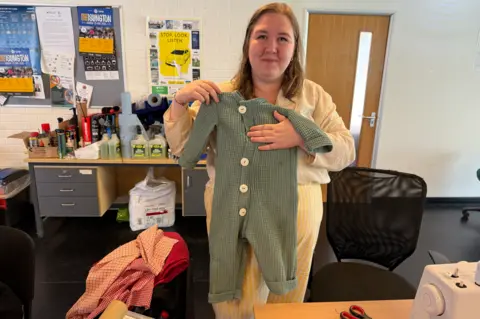 A women standing in an office holds a green baby grow which has white buttons down the middle of it