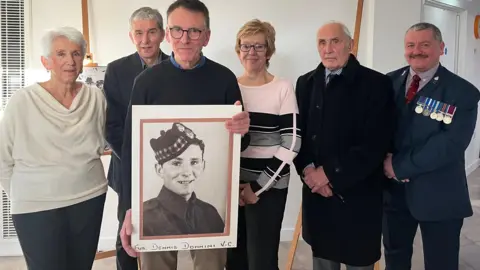 Group of six people posing for photo. Jerome Donnini is second from the right. There is also a man wearing a suit and medals on his breast-pocket. 
Two other women and three men, are dressed smartly. The man in the middle is holding a large photo of Dennis Donnini wearing a military hat. 