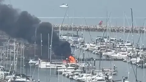 A picture of a fire, with thick black smoke pouring out of yachts, with boats moored around it in a marina.
