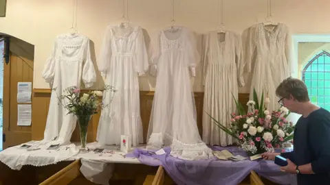 A selection of white dresses hanging up on a wall with flowers in front of them and a woman reading a card in front of them