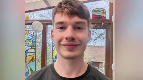 Edwin Thompson Photo of Edwin Thomas smiling to the camera. He is standing in front of stain glass window and is wearing a dark grey t-shirt.