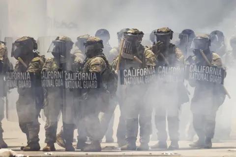 National Guard police walk through smoke holding up barriers.