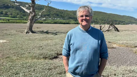 Jeff Cox is standing in the salt marsh with his hands in his pockets. In the background is the a green hill with trees.