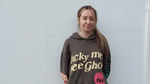 Henry Godfrey-Evans/BBC Abbie Parrish smiling with a brown hoody stood with her back to a white wall