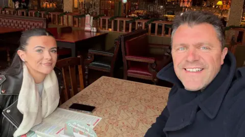 Alex is in a pub with his daughter Izzy. He is taking a selfie and wearing a heavy dark blue coat. She has he hair tied back and is wearing a black leather jacket with a cream and grey scarf.