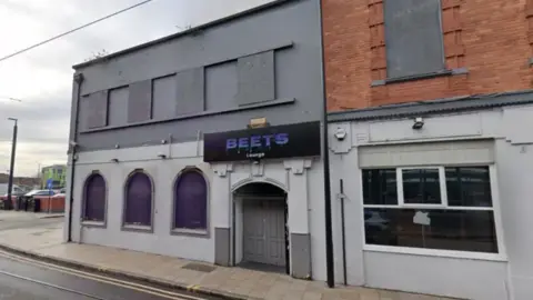 The building is painted grey with a purple/grey sign outside above the entrance. Upper and lower windows appear blocked out. It sits on the end of a street.