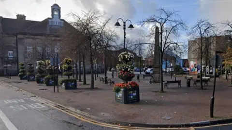 A view of a pedestrianised area of Leigh town centre. There are planters filled with brightly coloured flowers, benches and a war memorial.