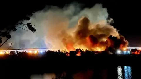 Flames and smoke rising in a night sky. A large expanse of water is in front of houses well alight.