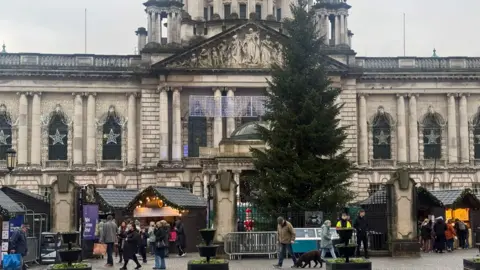 A wide shot of Belfast's Christmas tree outside City Hall. There are many small, wooden Christmas market huts around the tree as it stands outside a large, white stone building with pillars