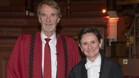 Sir Jim and Prof Tracey stood next to one another at the ceremony. Sir Jim is wearing a red tie and a red gown, Whilst Prof Tracey is wearing a white bow tie and a black gown.