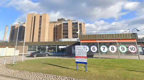 The main entrance to Raigmore Hospital. There is a sign on an area of grass and behind it the numbers and colours of different zones inside the hospital.