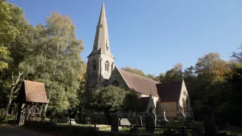 Laura Foster/BBC An old church with a steeple and surrounded by tombstones and graves is enclosed by an iron railing and stone wall. The church is surrounded by trees and the sky is blue, even though it is an autumnal day.