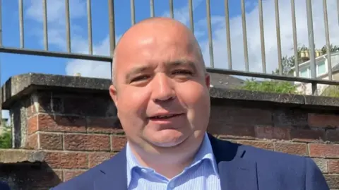 Brian Tierney is seen in a light blue and whiteshirt and navy blazer. He is standing in front of a red brick wall on top of which is metal railings.