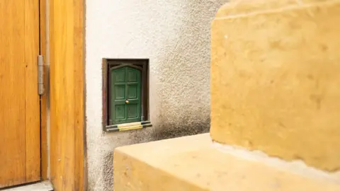 James Beck A miniature green wooden door hidden by the foot of a large Brown wooden door.