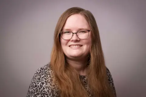 Emily with long reddish hair, parted in the middle and framing her face. She is smiling and wearning black rimmed glassed and a flower patterned black and grey top