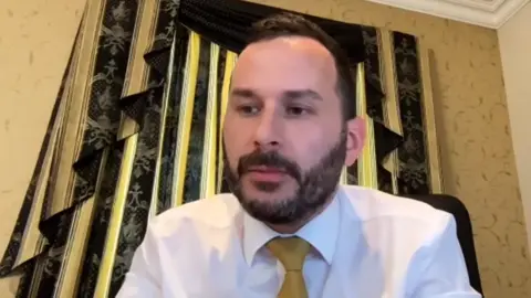 NI Assembly A man with black hair and beard wearing a white shirt and yellow tie sitting in a room with yellow wallpaper and in front of black curtain for a video call.