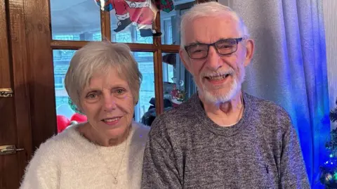 TamesideCouncil Sue and Pete Williams smile stood in a living room. Sue is wearing a white jumper while Peter is wearing a brown jumper.